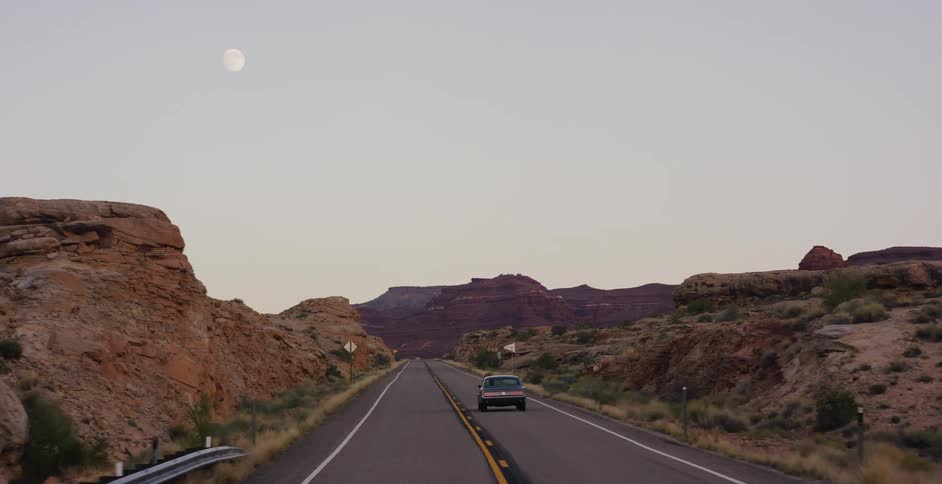 A car drives down a lonely desert highway through red rock canyons as a full moon rises in the evening sky