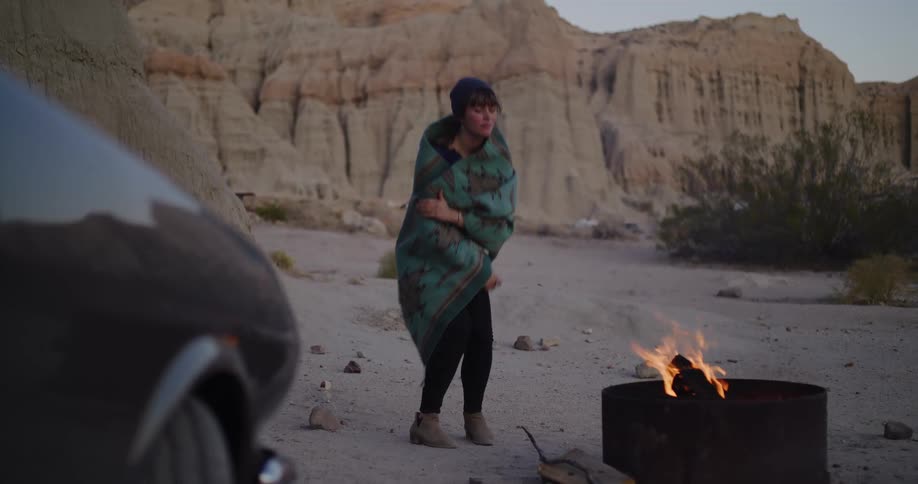 A woman wrapped in a blanket warms herself by a campfire while camping in a desert canyon at dusk