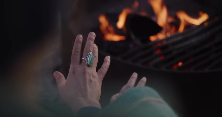A person wearing a large gemstone ring warms their hands by a crackling campfire