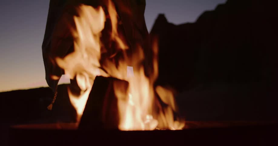 A solitary woman wrapped in a cozy blanket warms her hands over a crackling campfire at twilight as the camera slowly tilts down