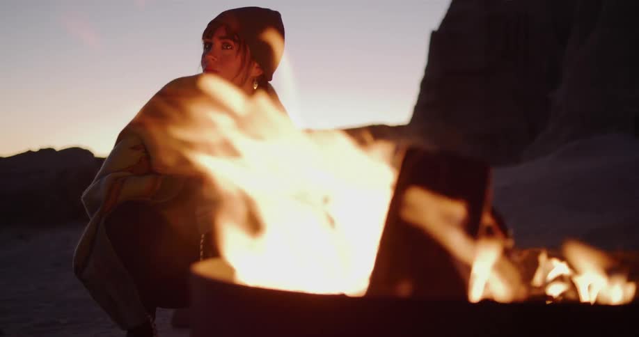 A woman wrapped in a blanket warms her hands by a crackling campfire at dusk as sparks fly in slow motion