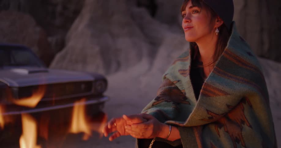 A woman wrapped in a blanket warms her hands by a glowing campfire in the desert at dusk, with her vintage car in the background