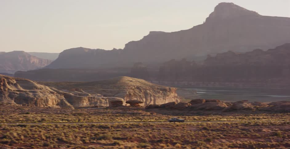 An aerial view follows a lone pickup truck driving along a desert highway surrounded by vast red rock canyons at golden hour
