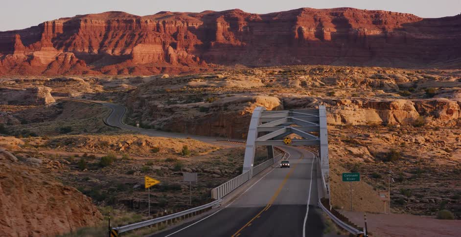 A classic black car drives across a long bridge spanning a vast desert canyon landscape during the golden hour