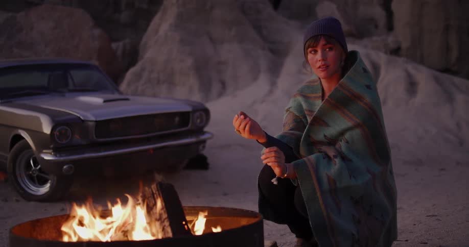 Cinematic slow motion shot of a woman wrapped in a cozy blanket warming herself by a campfire as sparks fly, with her classic car in the desert background