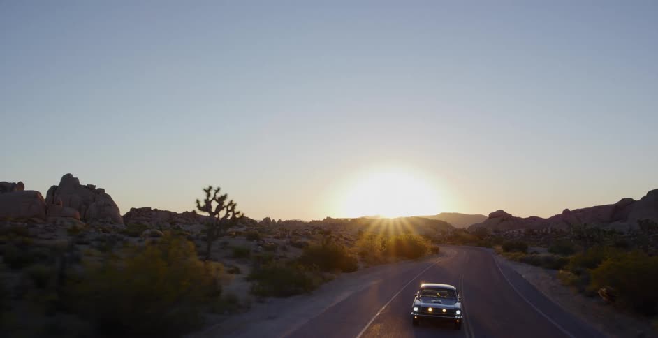 Aerial footage shows a vintage car driving along a winding desert road in Joshua Tree National Park towards the setting sun