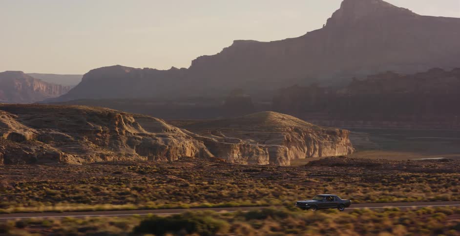 A classic muscle car drives down an empty highway through the vast desert landscape of the American Southwest at sunrise