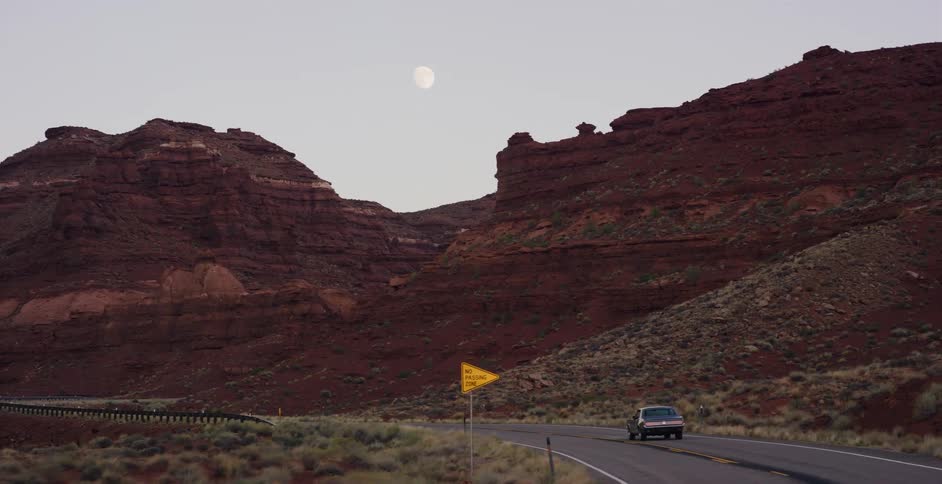 A classic car drives down a winding desert road through a red rock canyon under a full moon at dusk
