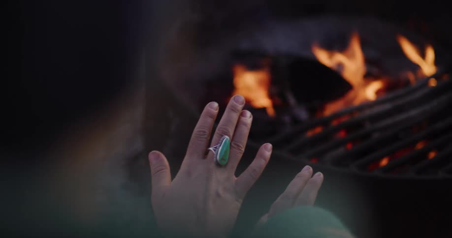 A person wearing a large turquoise ring warms their hands over a cozy campfire in slow motion