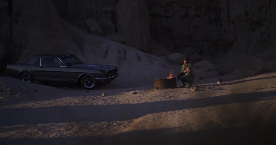 A lone traveler warms by a campfire next to a classic muscle car in a desolate desert canyon at twilight
