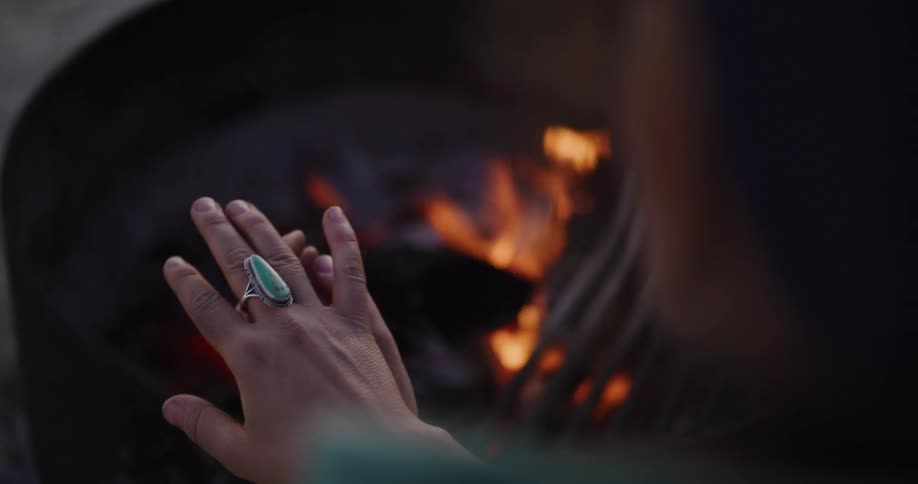 An over-the-shoulder view of a person with a turquoise ring warming their hands over a crackling campfire