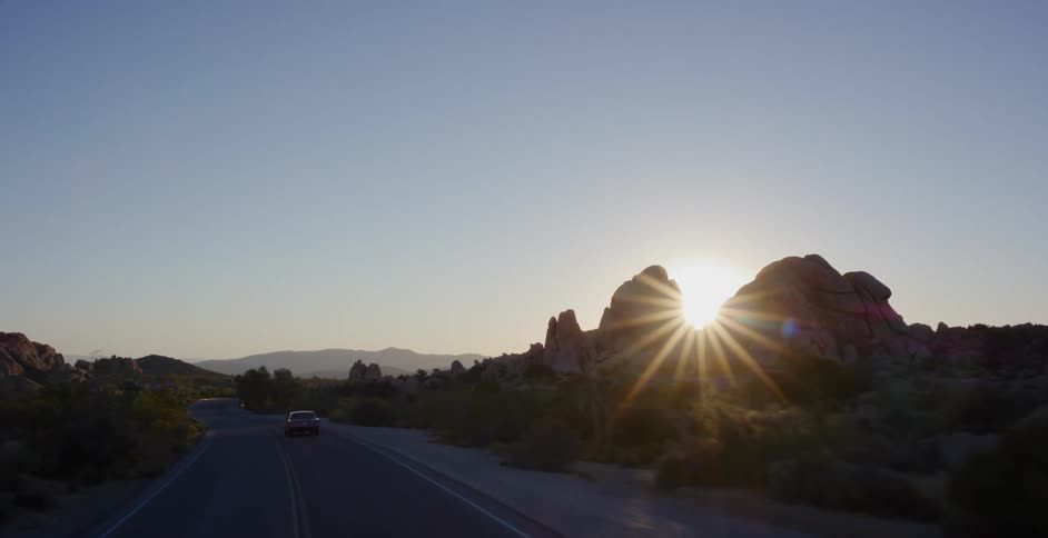 The sun bursts from behind a rocky silhouette as a car drives along a desert road at dusk