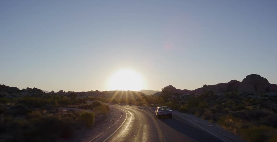An aerial view follows a vintage car driving along a winding desert road towards a brilliant sunset on the horizon