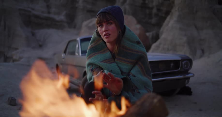 A woman wrapped in a blanket and wearing a beanie warms her hands by a campfire in a rocky desert landscape with a vintage car in the background