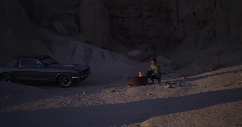 A solitary figure warms their hands by a campfire next to a vintage muscle car in a vast desert canyon at dusk