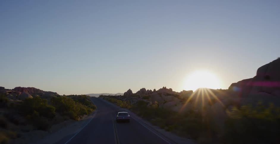 Aerial shot follows a vintage car driving along a scenic desert road into the sunrise
