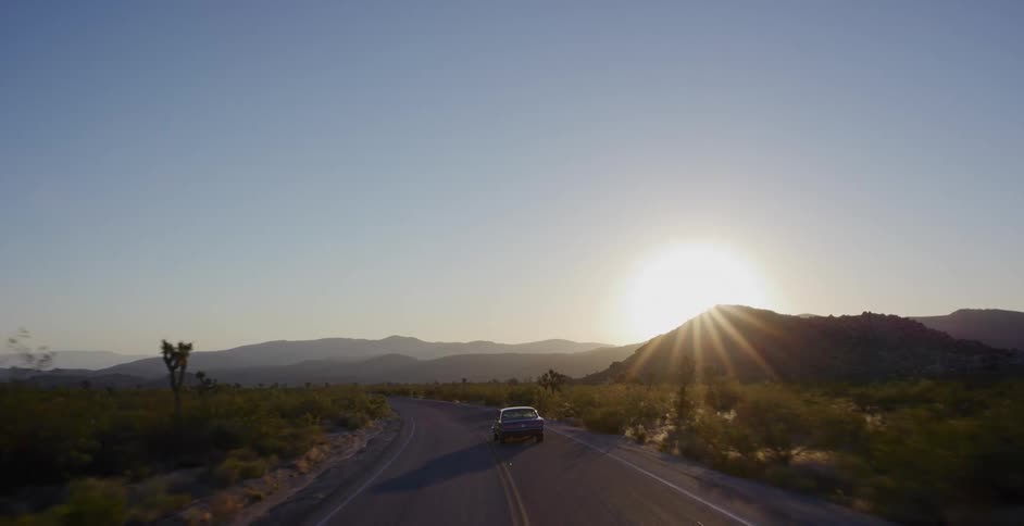 Aerial view of a car driving on a scenic desert road towards the setting sun over distant mountains