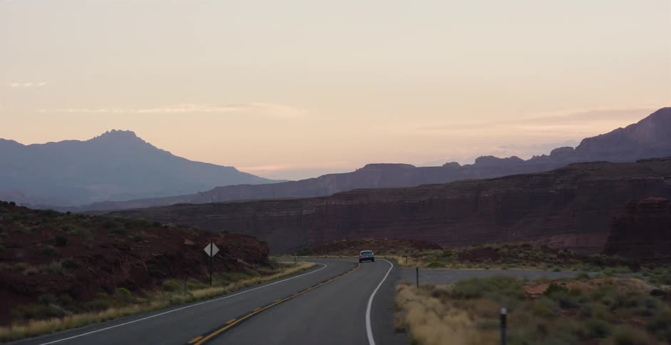 A lone car travels down a winding desert highway through majestic red rock canyons and mountains at dawn