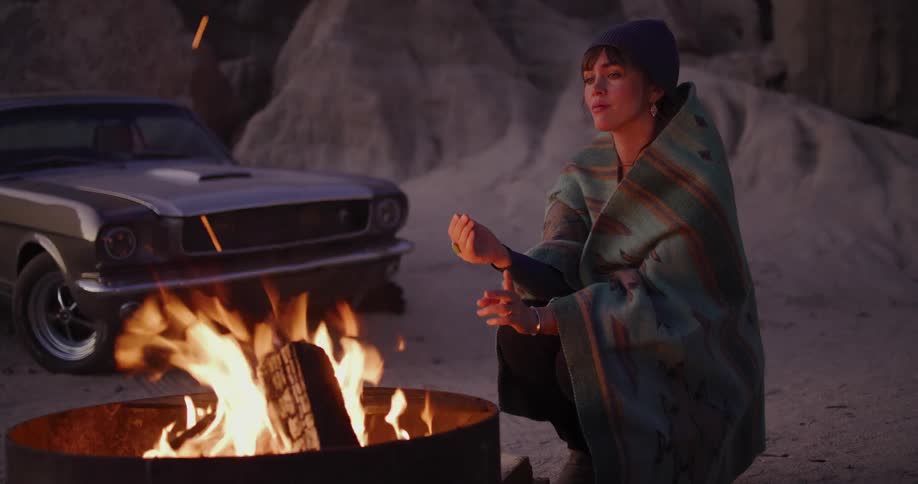 A young woman wrapped in a cozy blanket warms her hands by a crackling campfire in a desert canyon at dusk, with a classic muscle car in the background