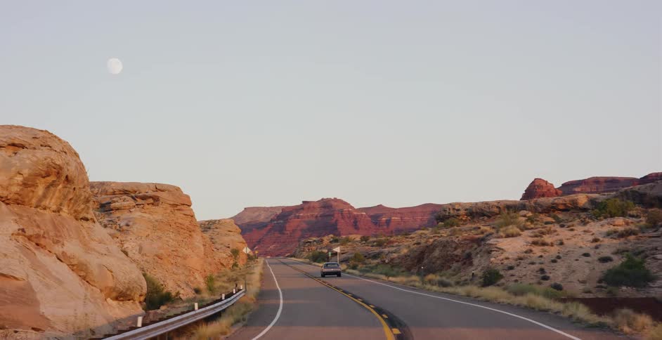 A car drives down a scenic highway through a red rock desert canyon at dusk as the full moon rises in the sky