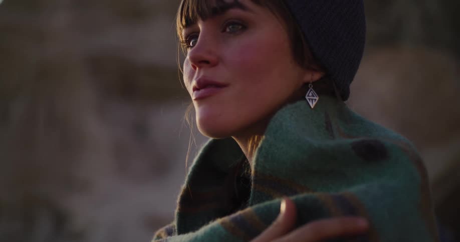 A serene young woman wearing a beanie and wrapped in a warm blanket gazes thoughtfully into the distance during golden hour