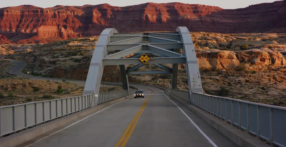 A cinematic tracking shot follows a vintage car driving across a bridge spanning a vast red rock desert canyon at golden hour