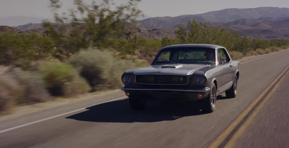 A classic silver Mustang cruises down a sun-drenched desert highway with mountains on the horizon