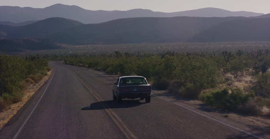 A classic black car drives down an empty two-lane highway through the desert with mountains on the horizon