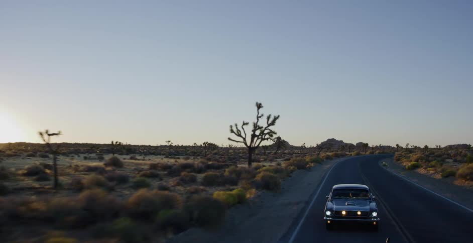 An aerial view of a classic muscle car driving down a scenic desert highway surrounded by Joshua trees at sunset
