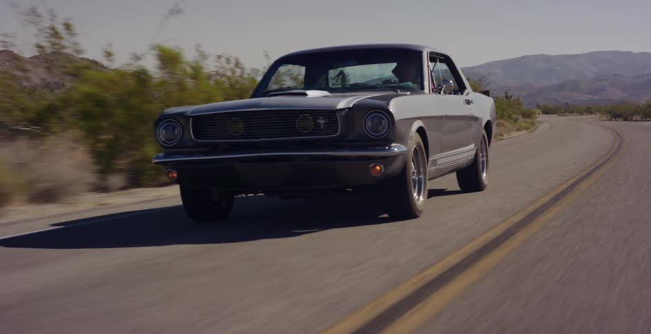 A classic grey Ford Mustang cruises along a winding desert highway on a sunny day