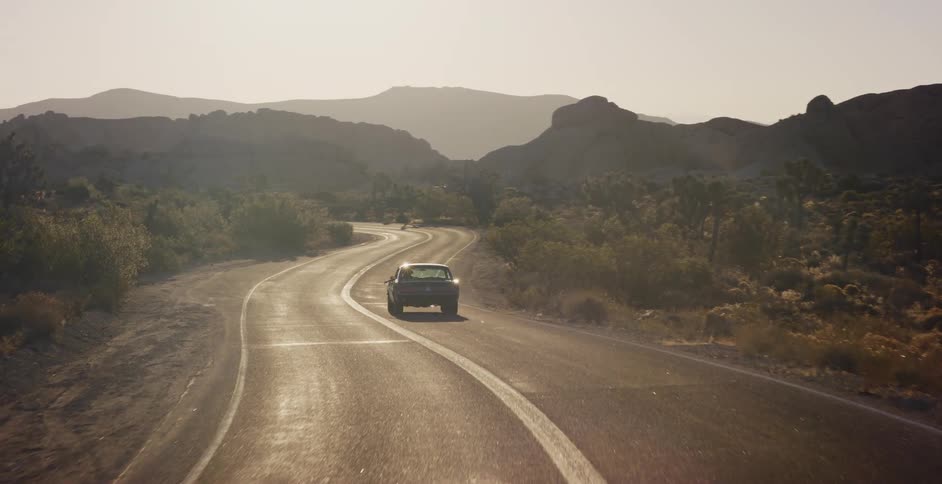 An aerial shot follows a classic car driving away on a scenic, winding road through a desert mountain landscape during a hazy sunset