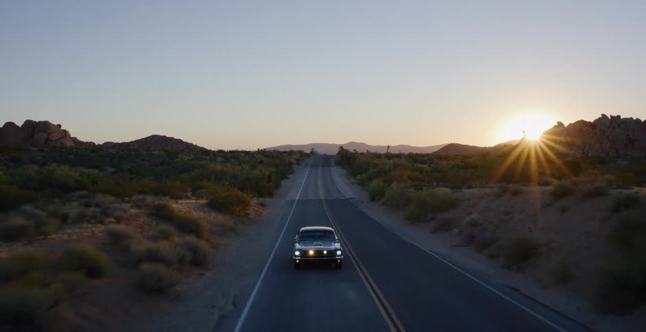 An aerial view captures a classic black car driving down an empty two-lane desert highway into the beautiful sunset