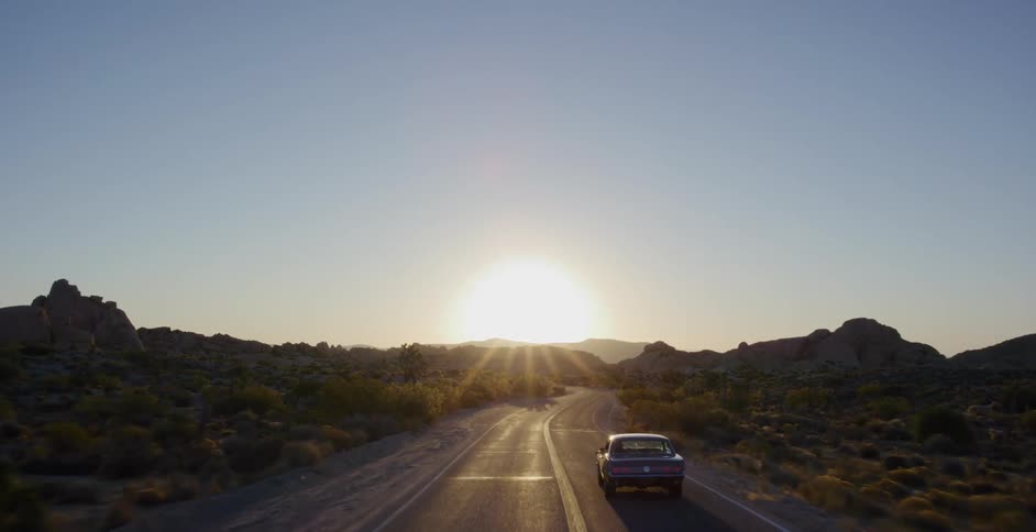 A classic muscle car drives along a scenic winding road through the desert towards the glowing sunset
