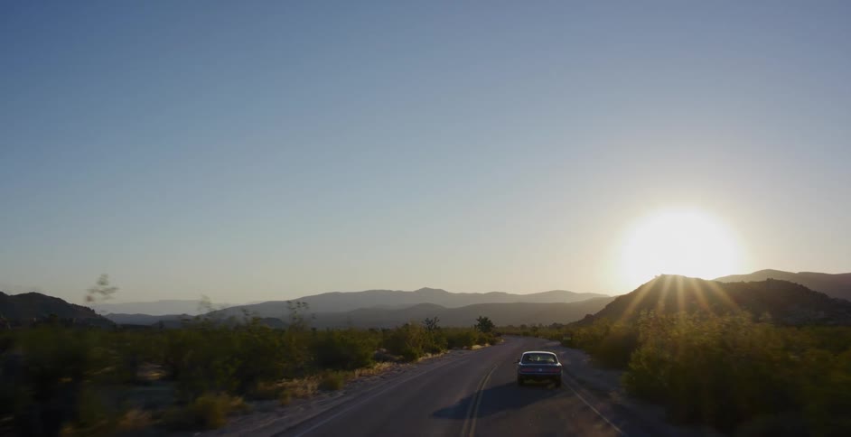 A classic car drives through the desert landscape of Joshua Tree National Park as the sun sets over the mountains