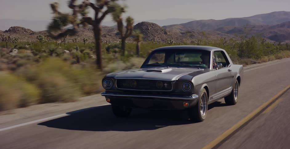 A classic silver Ford Mustang cruises down a two-lane highway through a sunny desert landscape with mountains in the background