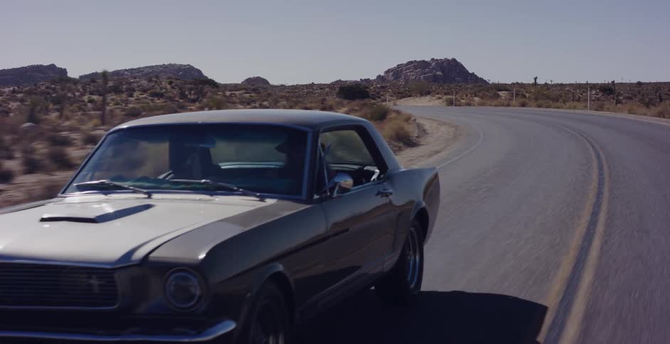 A classic silver Mustang coupe cruises along a winding desert road during a sunny daytime road trip