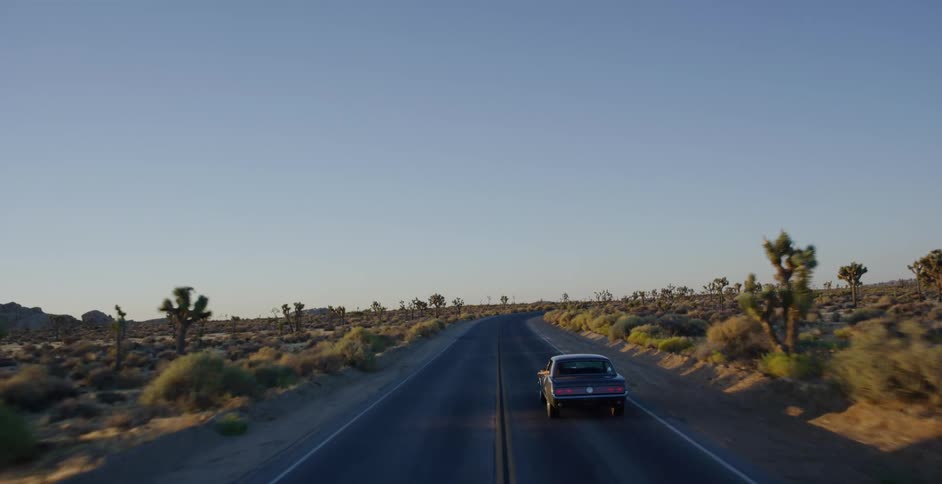 A drone follows a vintage muscle car driving down a remote desert road at sunset