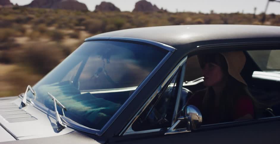 A free-spirited woman in a hat drives a classic car on a sunny road trip through the vast desert landscape