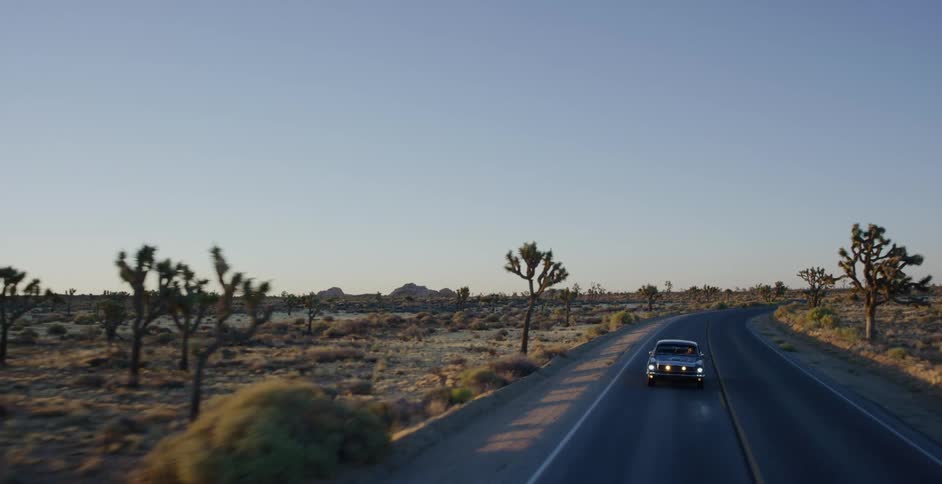 An aerial view shows a vintage car driving along a winding road through a desert landscape dotted with Joshua trees at golden hour