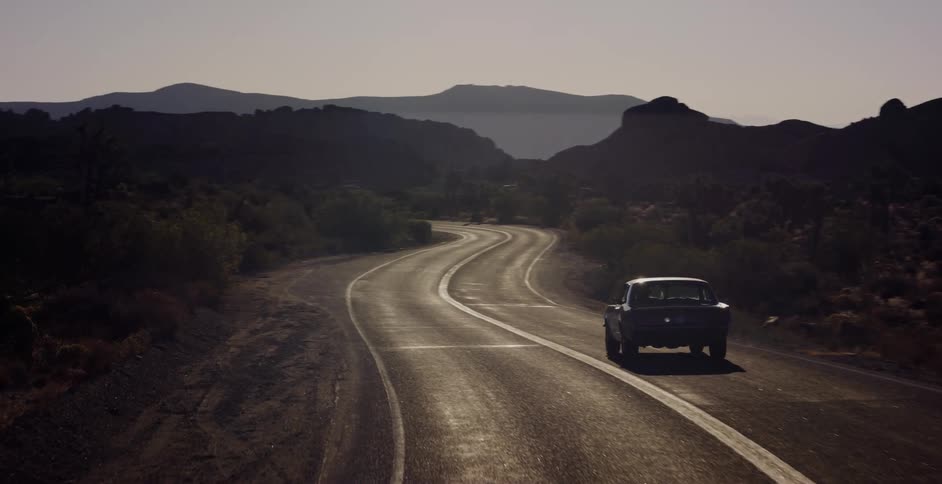 A classic black muscle car drives down a winding desert road at sunset, casting a long shadow