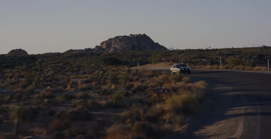 A classic American muscle car drives around a winding road through the scenic desert landscape at golden hour