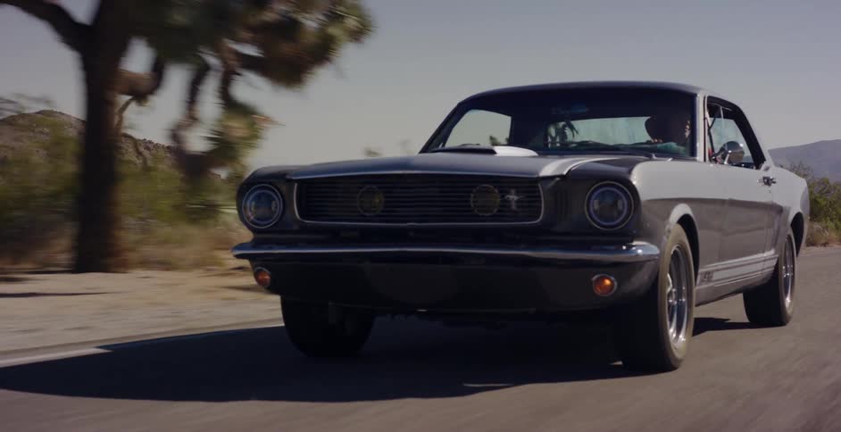 A classic Ford Mustang with racing stripes drives along a desert highway past Joshua trees on a sunny day
