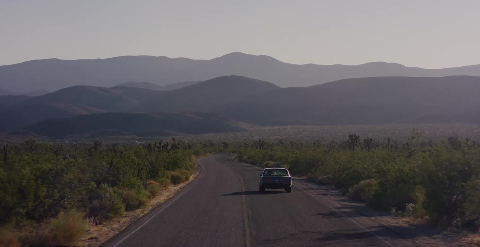 A classic car drives along a winding road through a vast desert landscape towards hazy mountains in the distance