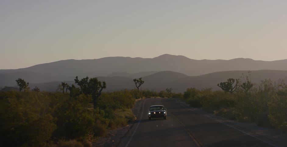 A vintage black car with headlights on drives down a remote desert road at dusk with Joshua trees and hazy mountains in the background