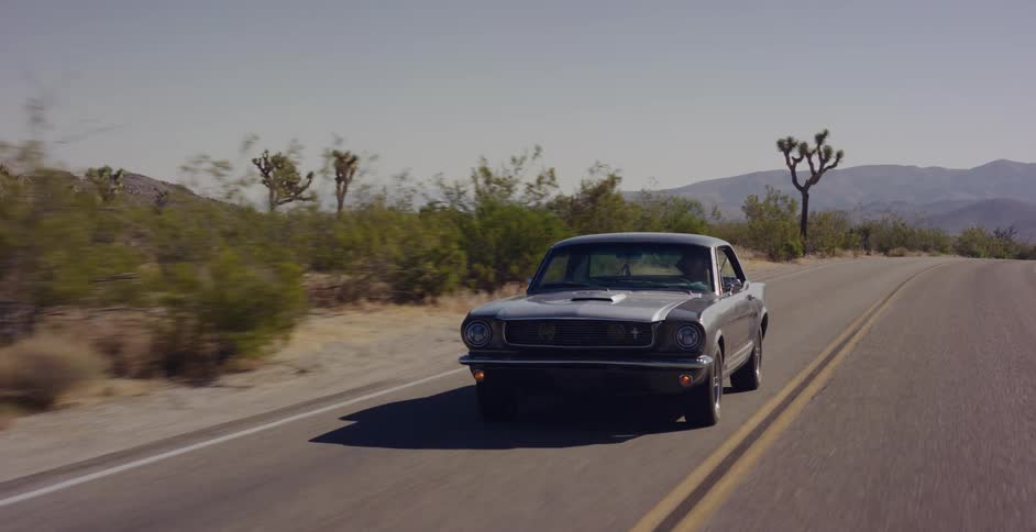 A classic silver Ford Mustang cruises down a scenic desert highway lined with Joshua trees on a bright, sunny day