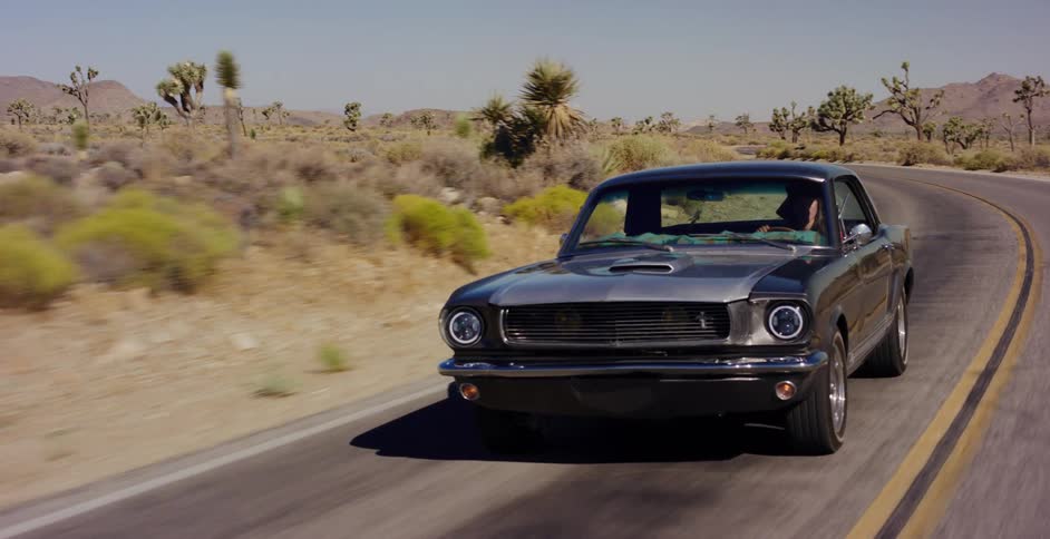A classic muscle car cruises along a winding road through the sunny desert landscape dotted with Joshua trees