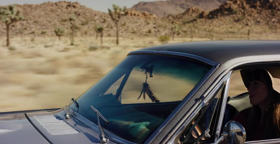 A woman enjoys a scenic drive in her classic muscle car through a desert landscape with rocky mountains and Joshua trees