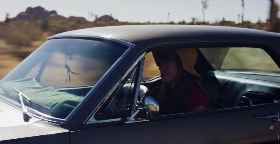 A smiling woman in a floppy hat enjoys a sunny, slow-motion drive through the desert in her classic car