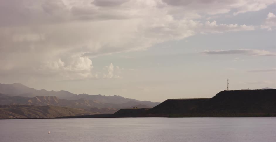 A serene, wide view of a calm reservoir with rolling hills and distant mountains under a cloudy sky