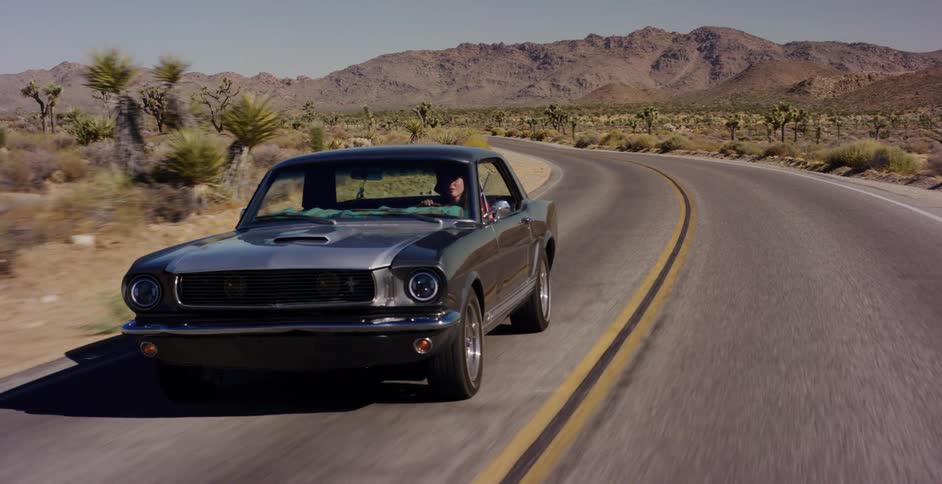 A vintage grey Ford Mustang cruises down a winding desert road surrounded by Joshua trees and mountains